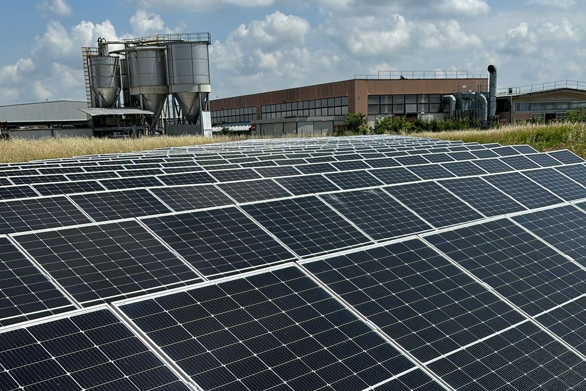 File di pannelli fotovoltaici a terra su erba secca, sullo sfondo capannone industriale con silos e cielo nuvoloso