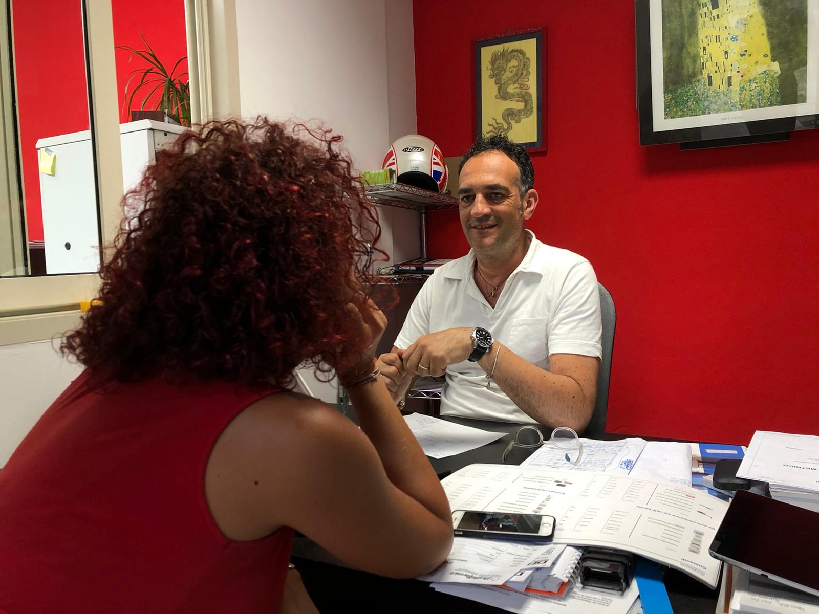Uomo in polo bianca alla scrivania piena di fogli parla con una donna dai capelli ricci ramati, pareti rosse e quadri